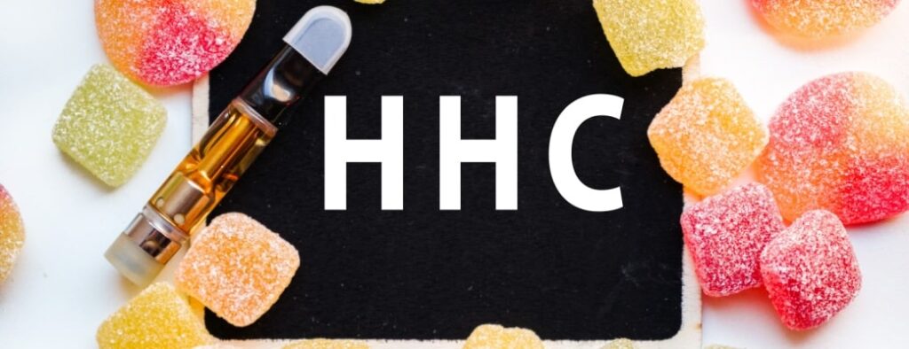 A small blackboard with the word “HHC” written on it is surrounded by gummy edibles and a vape cart. 